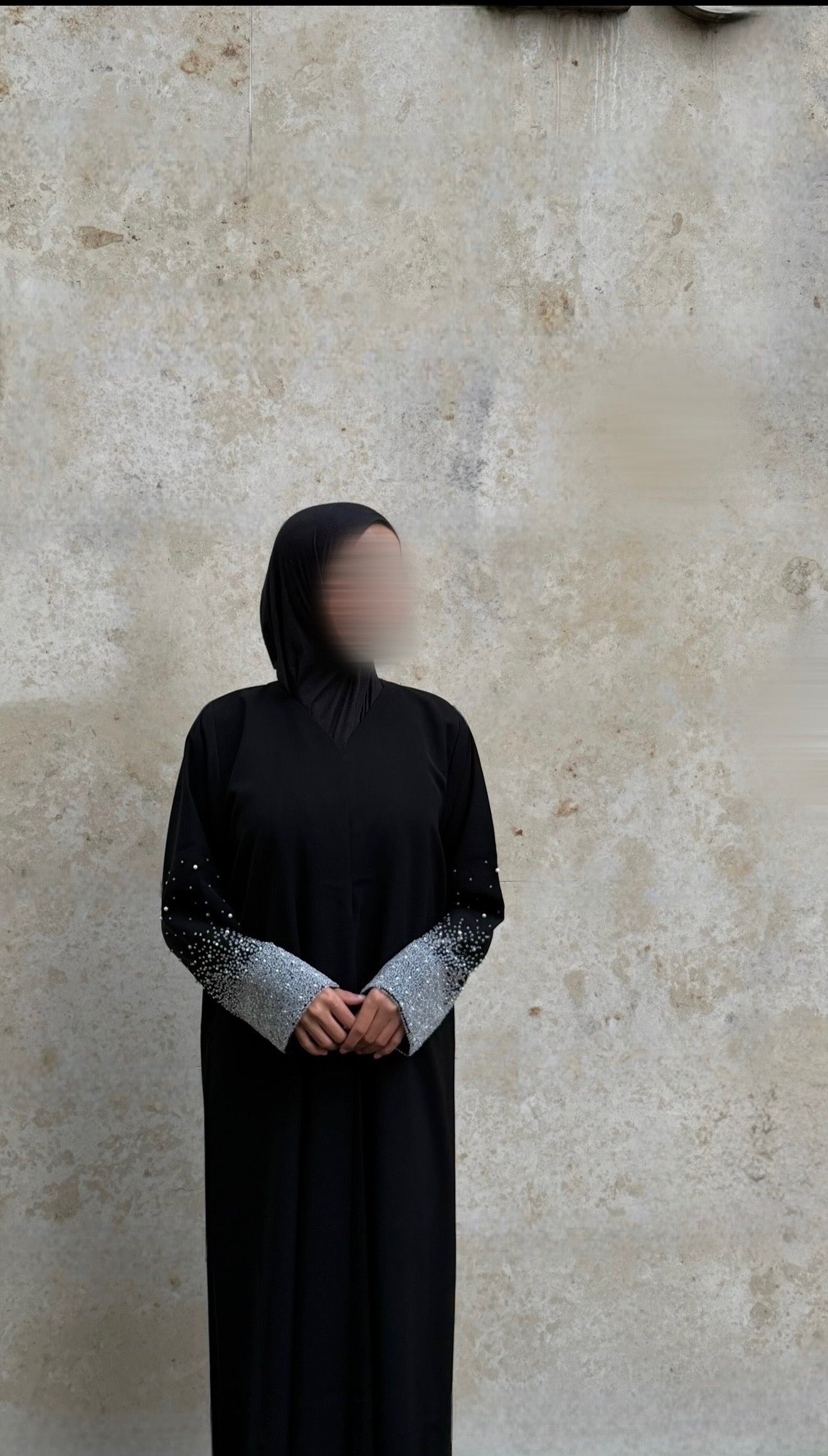 Person wearing a black abaya with silver embellishments against a beige wall.