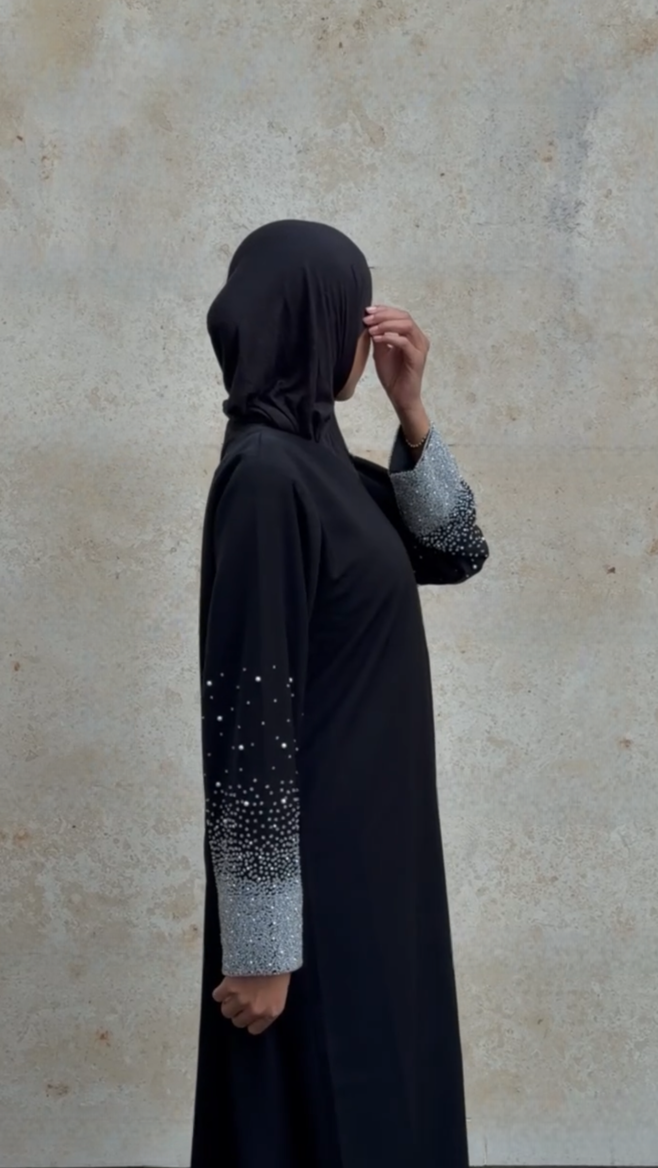 Person wearing a black abaya with silver embellishments against a beige wall.